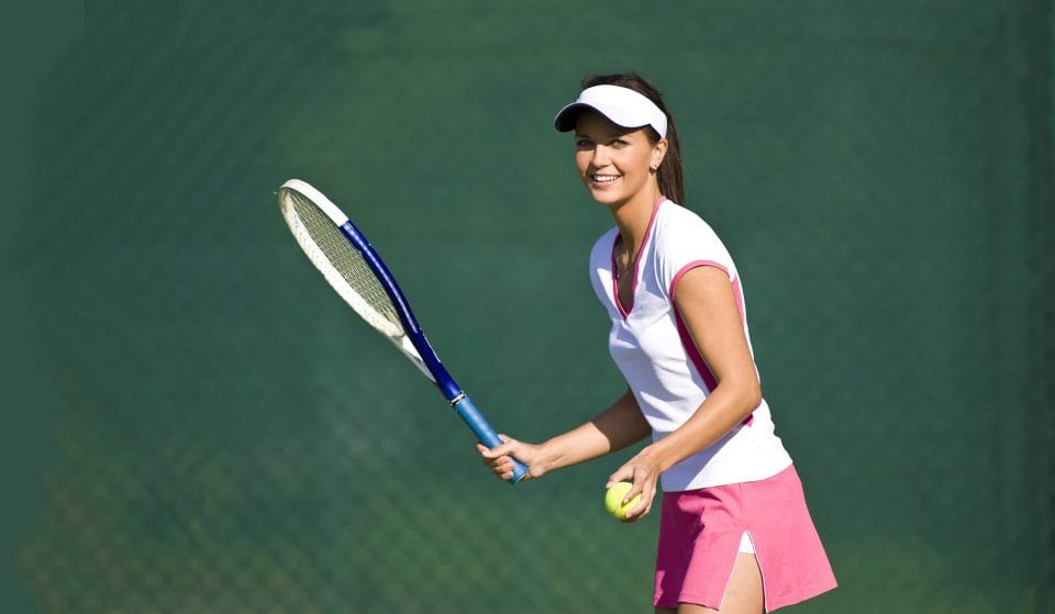 woman holding tennis ball and racquet in pink skirt and white shirt smiling on outdoor tennis court