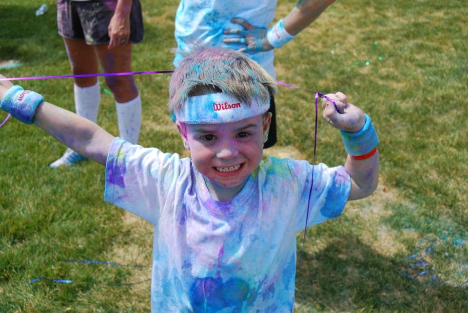 little boy with balloon string and smiling covered in bright blue, green, and purple during color run for kids
