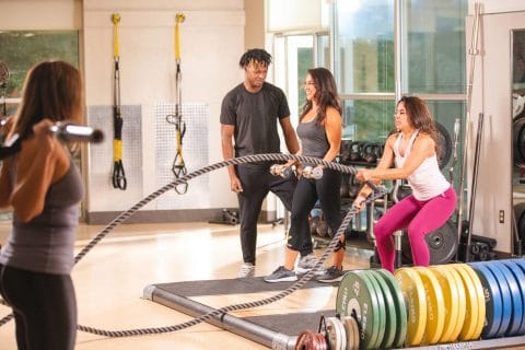 personal trainertraining woman with dumbbells while two other women workout on battle ropes and with a barbell