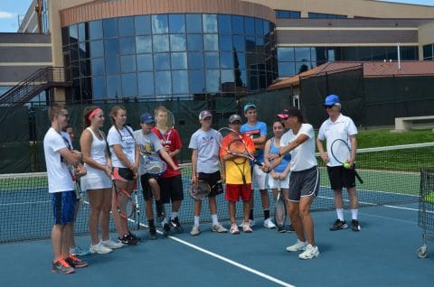 teens with tennis racquet smiling as they watch their coach demonstrate and shot