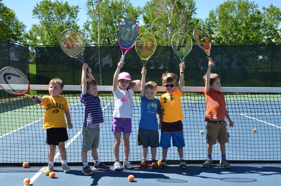 six small kids holding bright and colorful tennis racquets in the air and smiling on club greenwood outdoor tennis court