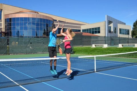 man and woman jump up to high five after tennis match on club greenwood outdoor courts with facility in the background