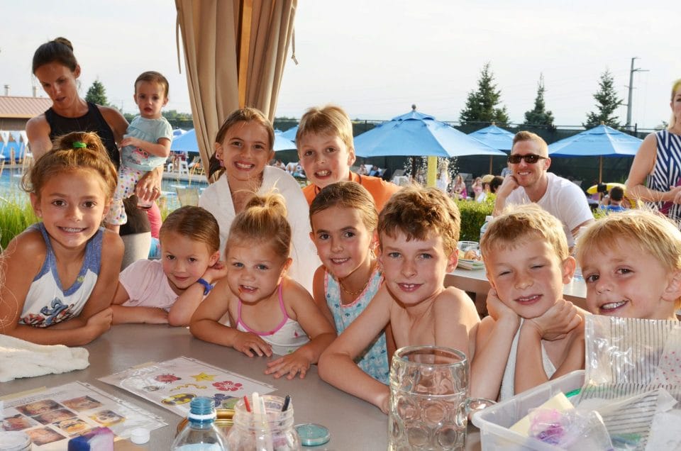 kids smiling at a table next to the pool with parents and blue umbrellas in the background