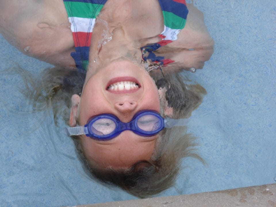 girl doing a backfloat and wearing goggles in outdoor pool for summer swim camp