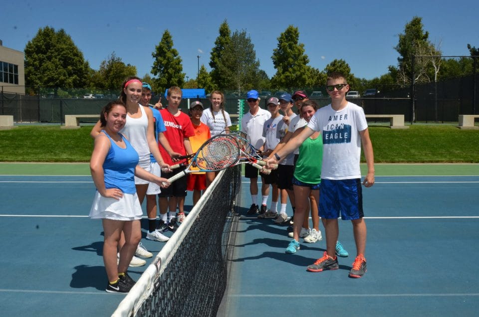 teens standing with tennis racquet next to tennis net and smiling