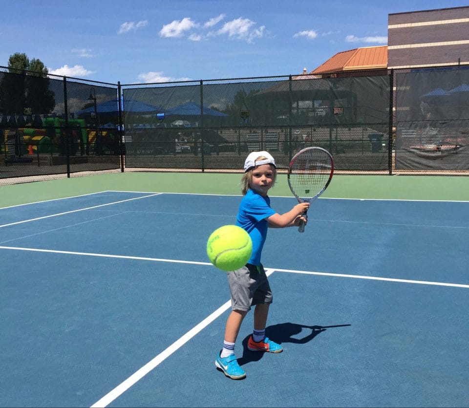 little boy in blue shirt and white hat getting ready to hit a tennis ball with his racquet on outdoor court