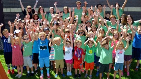 Big group of kids and camp counselors smiling during summer camp with hands up
