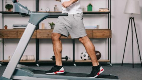 man walking on inclined treadmill at home wearing grey shorts a white shirt and black red and white running shoes