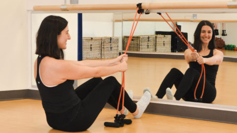 woman with dark hair is sitting on the floor in front of a mirror holding straps for a barre workout