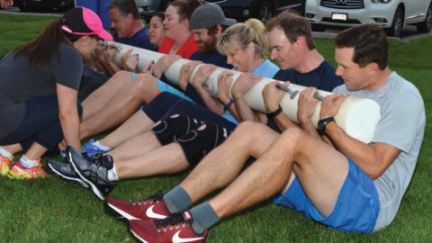 group of people working out holding a log and doing a log roll sit up outside
