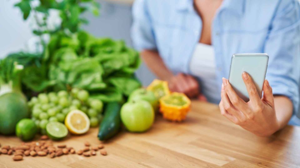woman holding phone next to a wooden table with fruits, veggies and nuts