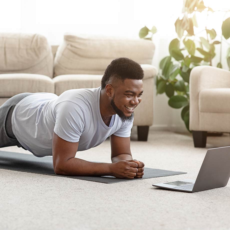 young man doing a plank on a mat at home smiling at a computer screen