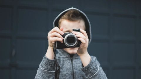 young boy wearing a gray hoodie taking a picture with a camera in front of a dark background