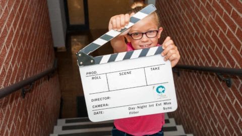 Young girl wearing pink shirt and glasses holding a clap board for making films