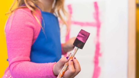 little girl wearing a pink shirt and blue smock painting with a brush and pink paint