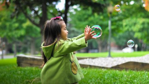 little girl in green jacket playing with bubbles outside in a park