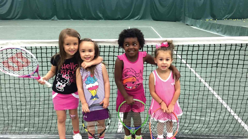 little girls on a clay tennis court with racquets smiling for the camera