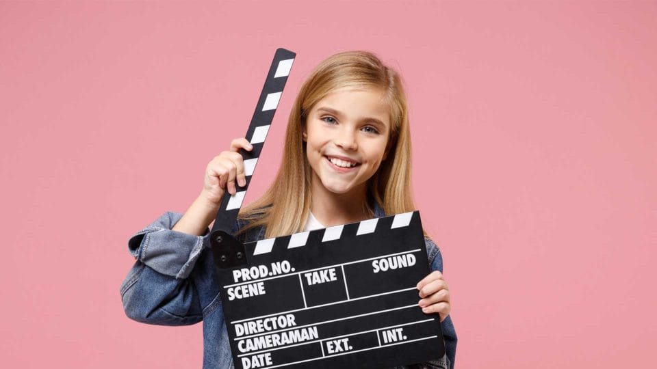 Young girl wearing a jean jacket holding a clap board for making films in front of a pink background